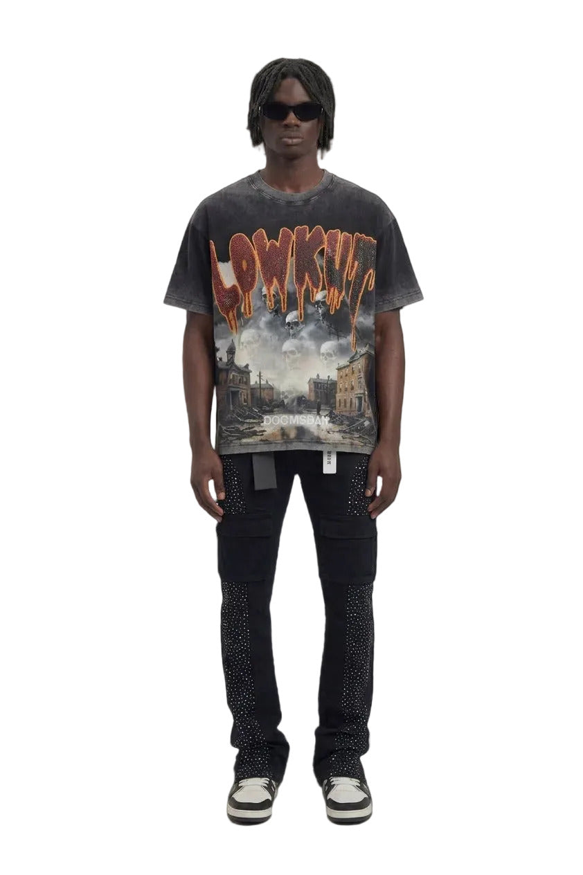 A model stands against a light gray studio background wearing a faded black graphic t-shirt with a distressed orange and white 'LOWKUT' logo and a cityscape print, paired with dark jeans and black-and-white sneakers.